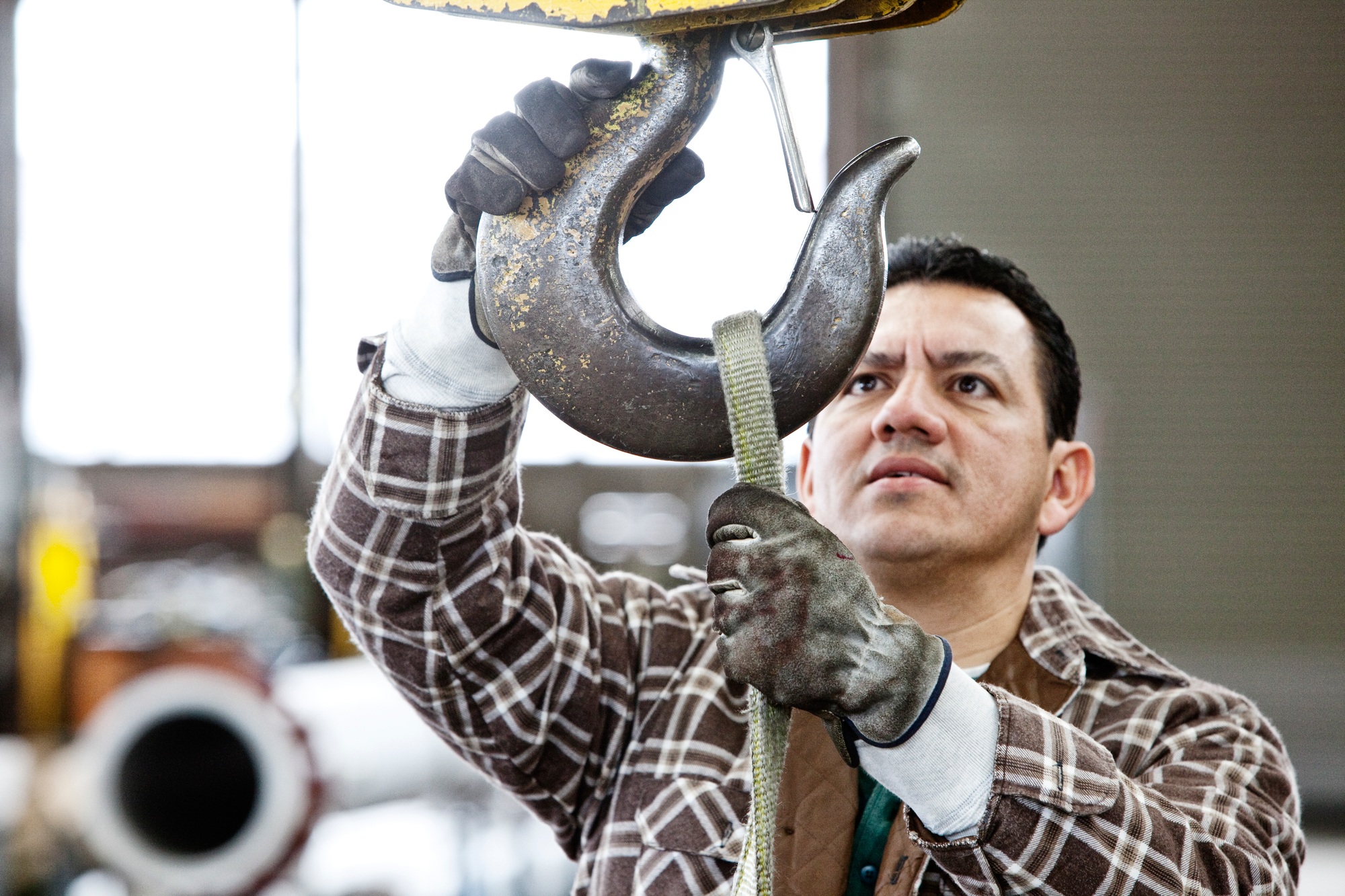 Hispanic man factory worker attaching a strap to the hook of a heavy duty overhead lift in a sheet